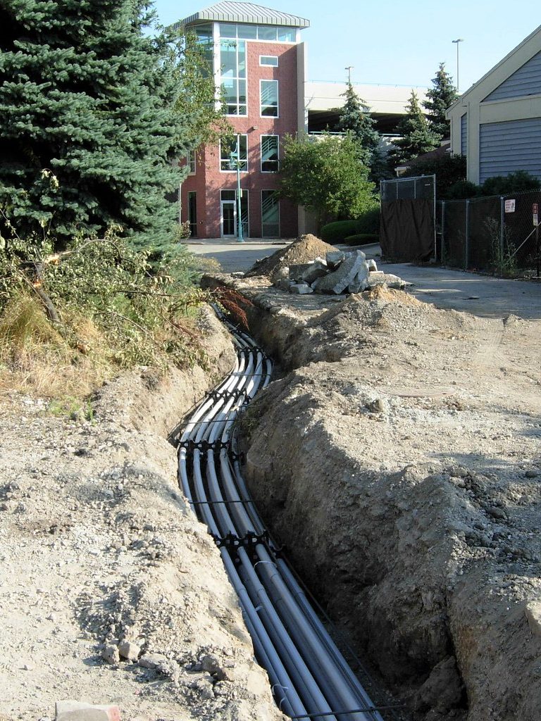 A long open trench running toward a building, carrying a bank of conduits held apart on spacers along its full length