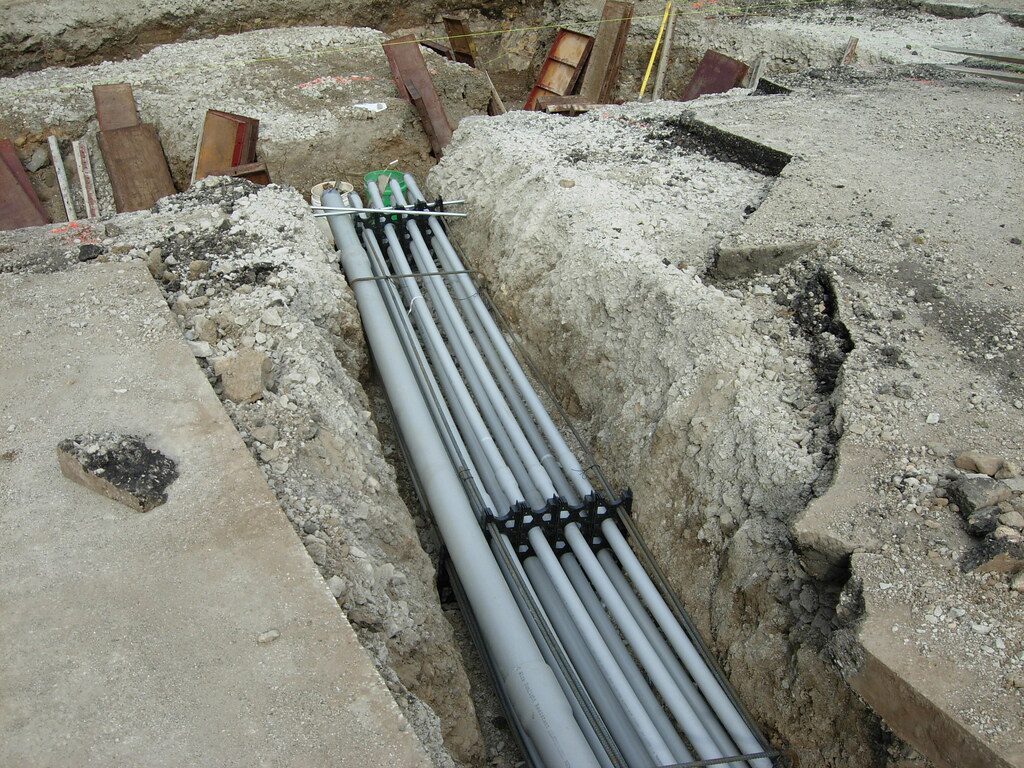 A bank of grey PVC conduits held on plastic spacers in an open trench, tied to rebar and ready for concrete encasement