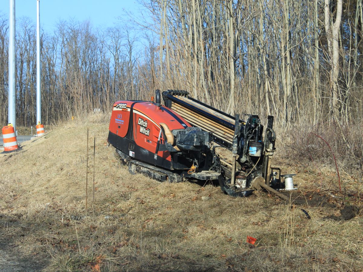 A tracked horizontal directional drill set up on a highway shoulder with a rack of drill rod loaded, an orange locate flag planted beside the entry point