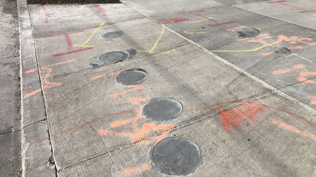 A concrete sidewalk with a diagonal row of six patched vacuum-excavation test holes, surrounded by orange, red and yellow utility locate paint marks