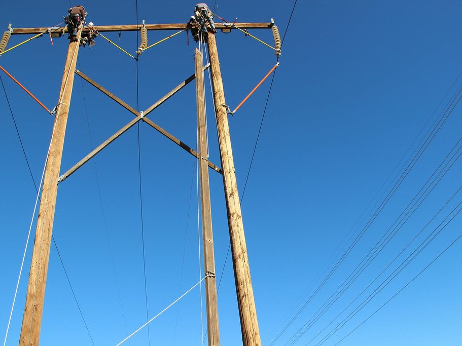 Two line workers at the top of a wood H-frame structure changing out a crossarm, conductors sweeping away against a clear sky